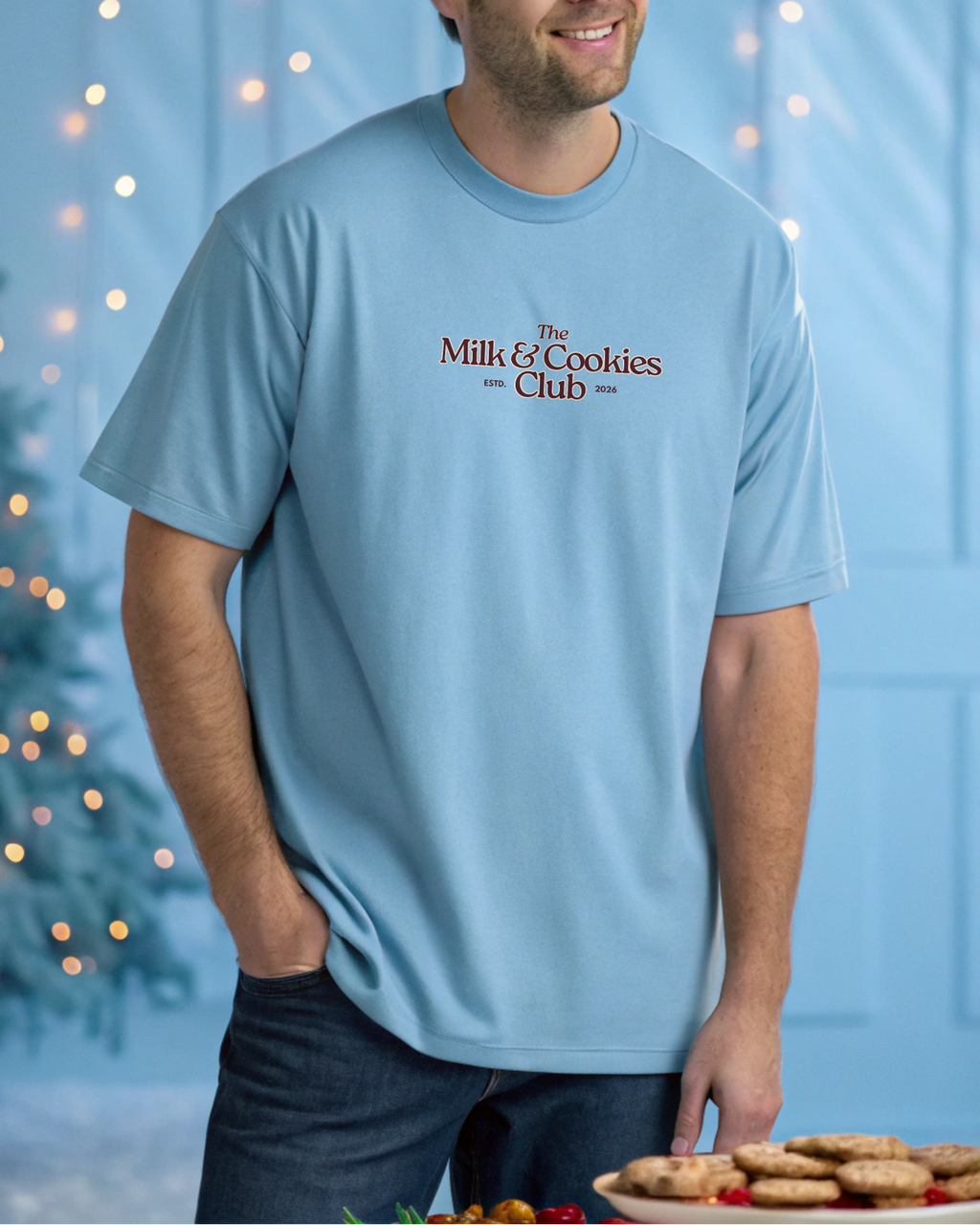 Man wearing a light blue t-shirt with 'The Milk & Cookies Club' text, standing in front of a Christmas tree with lights.