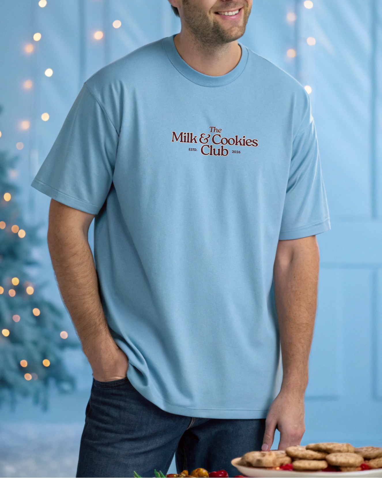 Man wearing a light blue t-shirt with 'The Milk & Cookies Club' text, standing in front of a Christmas tree with lights.